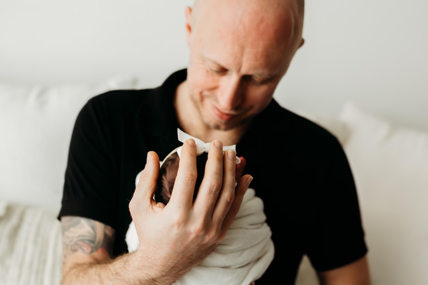A man in a black polo shirt holding a tiny newborn baby swaddled in a cream coloured wrap