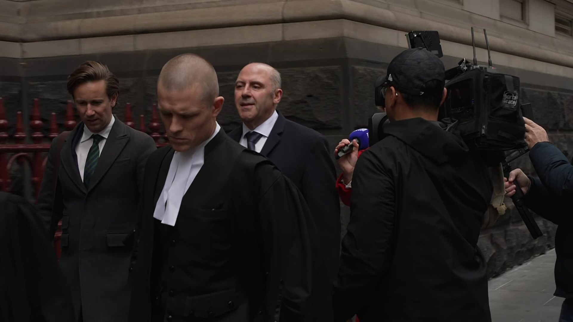 A balding man in a dark suit walks beside a stone building surrounded by people in black and tv cameras.