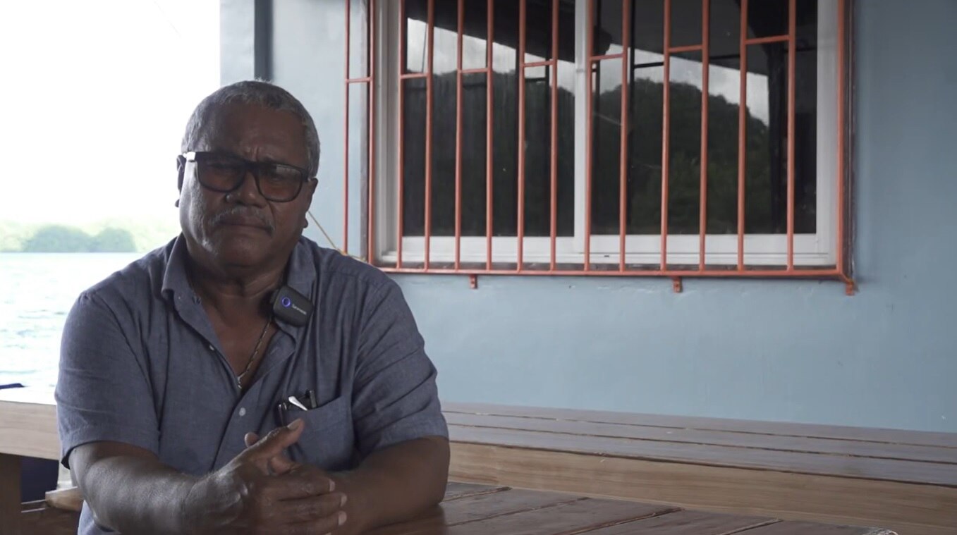 Natus Misech sits outside his office, with the sea in the background.