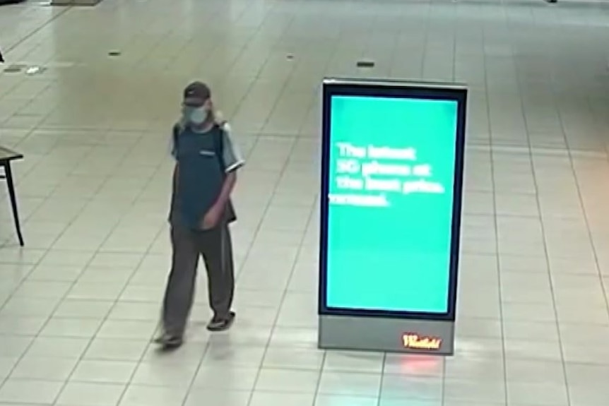 A man wearing a face mask and cap walks through a shopping centre next to an information screen
