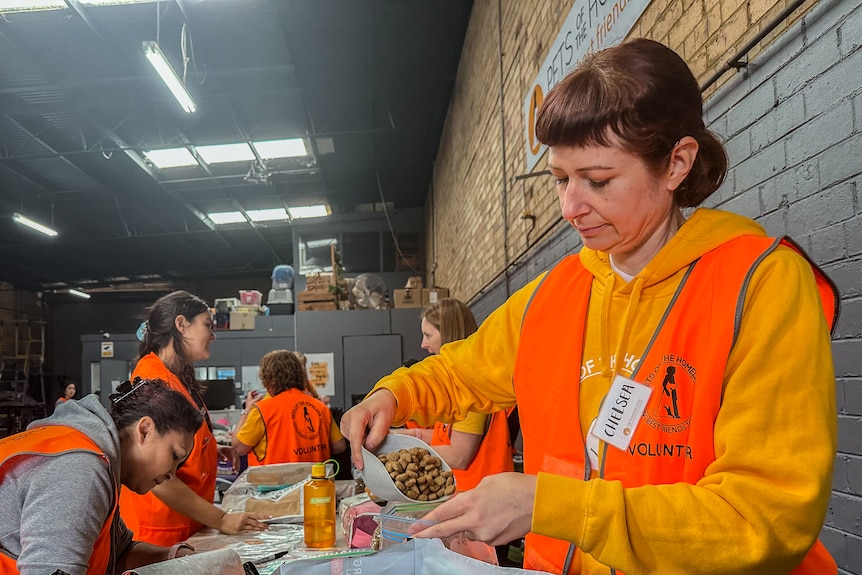 A woman in an orange jumper and vest scoops pet food into a bag, in a warehouse full of other volunteers.