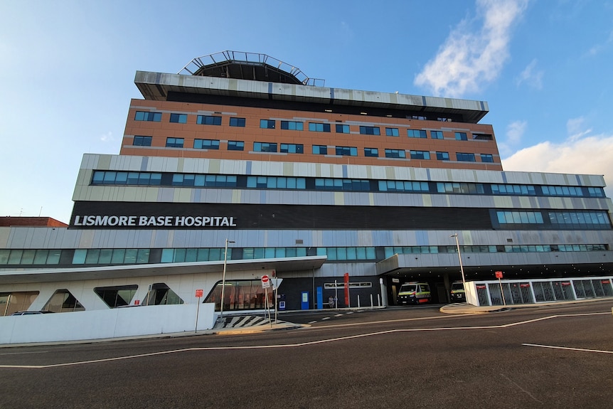 The exterior of Lismore Base Hospital with a helipad on top of a multi-storey building and ambulances parked at street level.