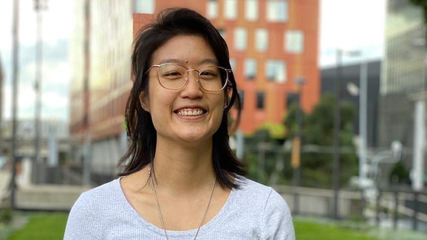 An Asian woman wearing glasses stands in front of a tall orange building with windows.