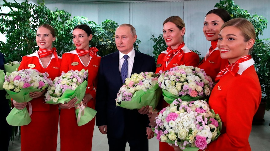 Vladimir Putin looking solemn while surrounded by young female flight attendants