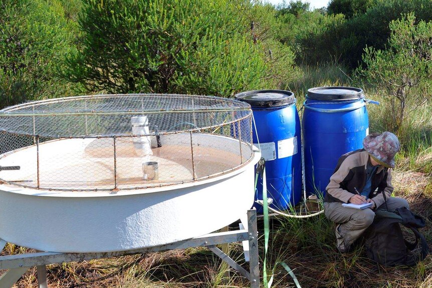 A woman crouches next to large drums and other scientific devices in bushland, taking notes.