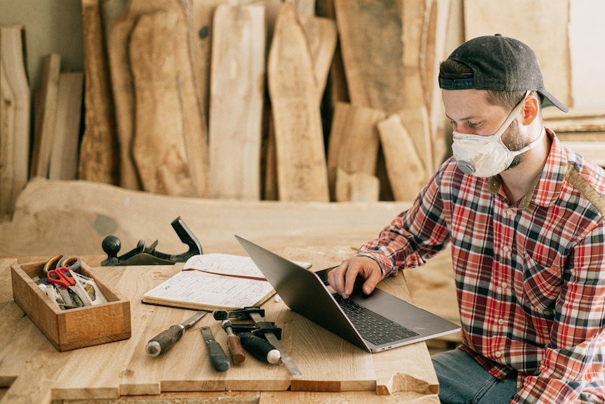 Man with a mask on works at a laptop against a backdrop of wood