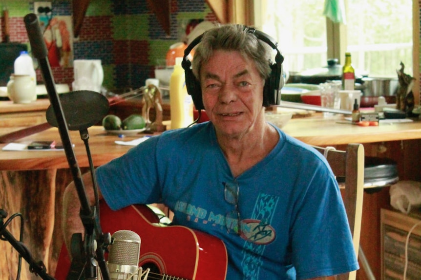 A man wearing headphones and holding a guitar sits in front of a microphone.