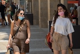 Two women walk down a city street carrying bags and wearing face masks 