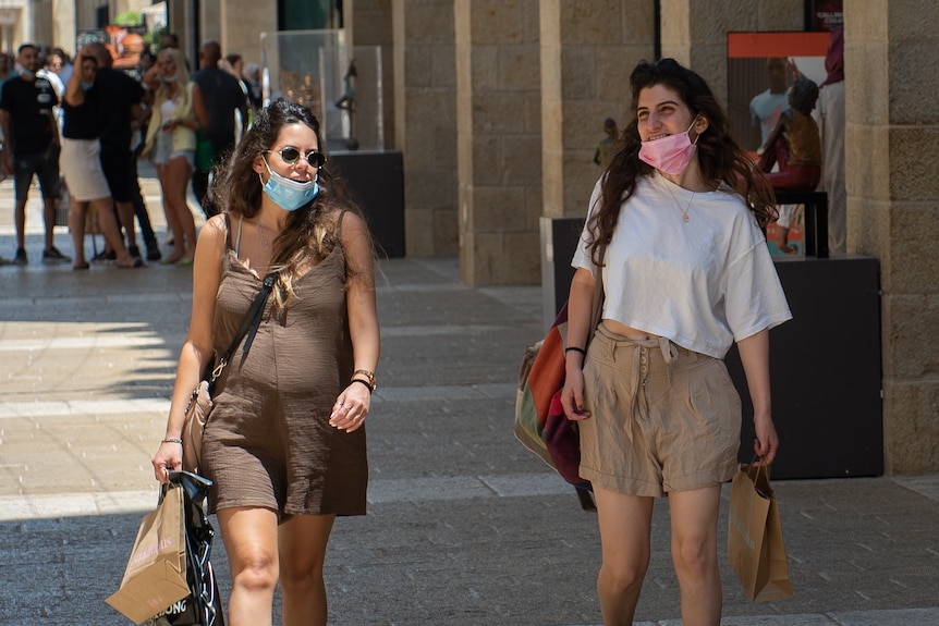 Two women walk down a city street carrying bags and wearing face masks 