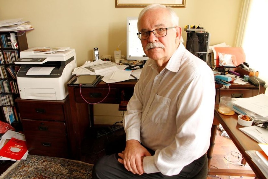 An elderly man with glasses in his office. He looks wise and concerned.