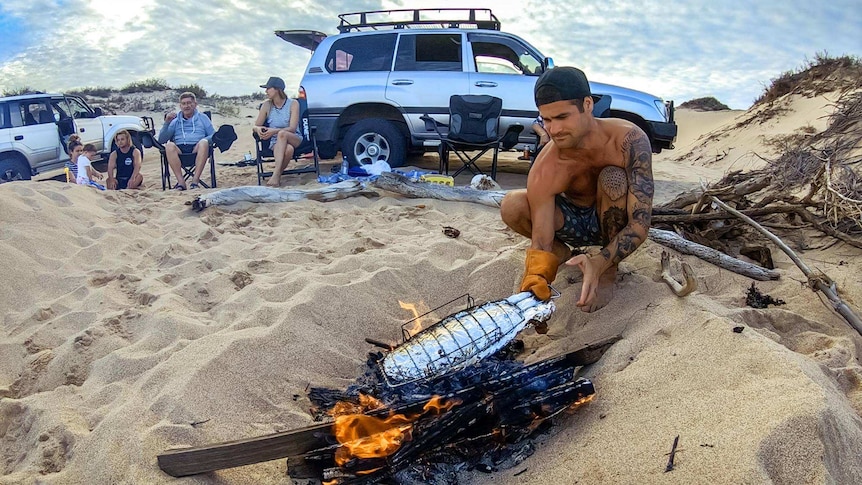 Tattooed, bare-chested man cooks fish wrapped in tinfoil over a beach fire.
