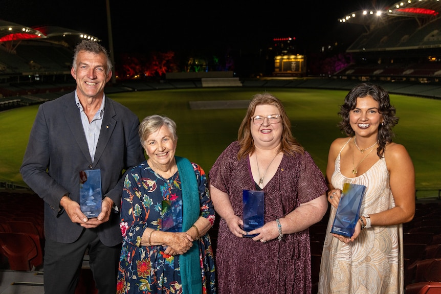 A man and three women posing with their awards in front of an oval