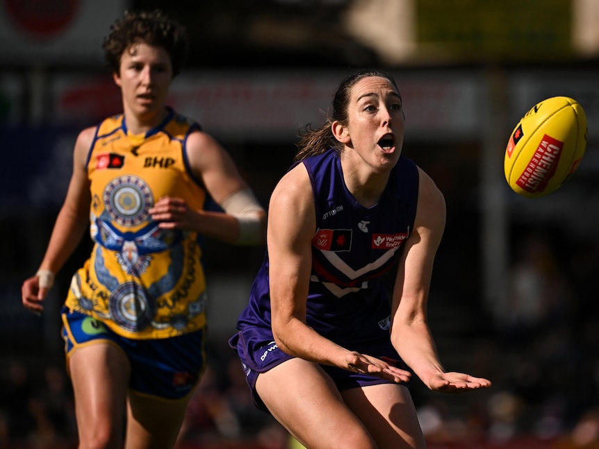 A Fremantle Dockers AFLW player reaches out low in front of her to grab the ball, with a defender trailing.