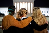 Three women stand with their arms around each other looking towards the Supreme Court, lit up in the night.