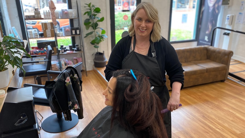 Woman standing at a hairdressing seat with a client, smiling. 