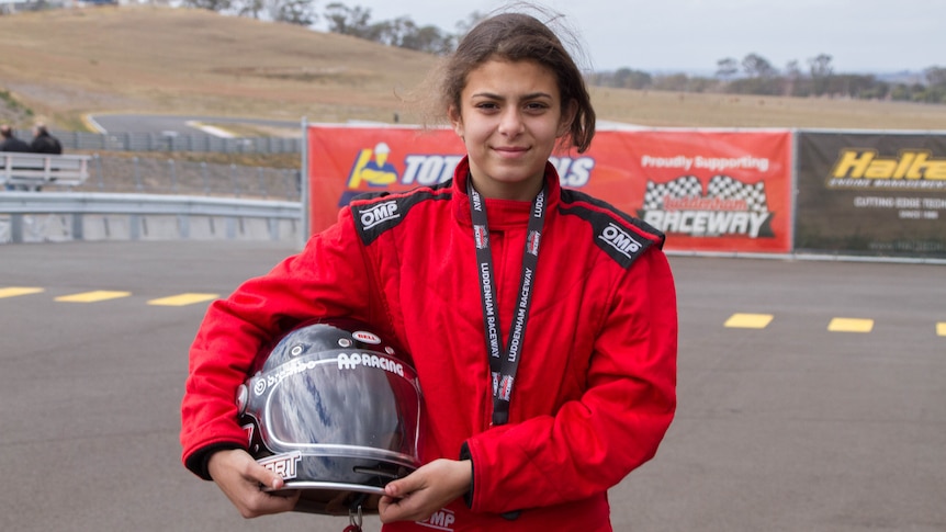 A girl stands holding a racing helmet by her hip wearing red racing overalls.