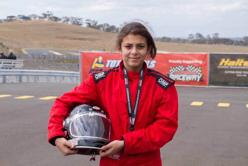 A girl stands holding a racing helmet by her hip wearing red racing overalls.