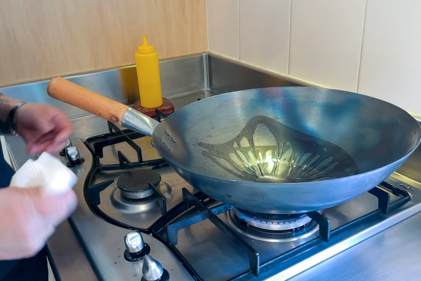 A wok on stovetop flame with oil drizzled in. Nearby a hand holding a paper towel.