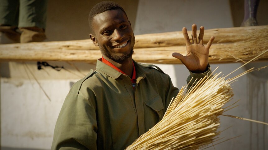 A man smiles and waves at the camera as he holds a stack of straw, outdoors