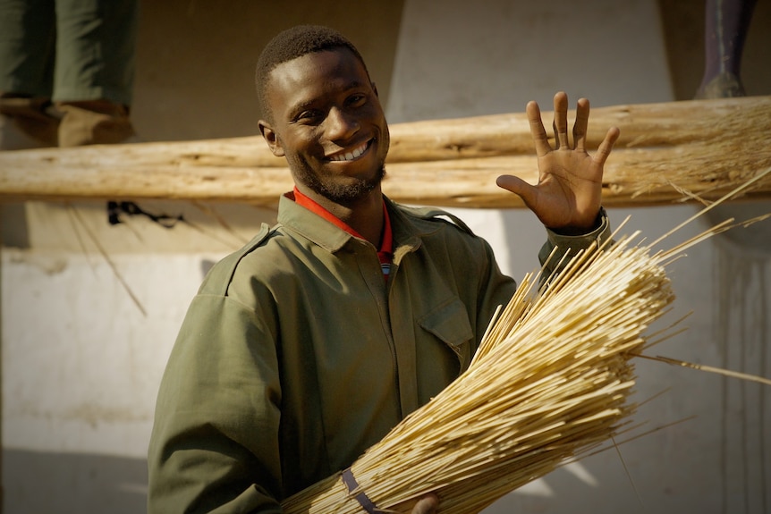 A man smiles and waves at the camera as he holds a stack of straw, outdoors