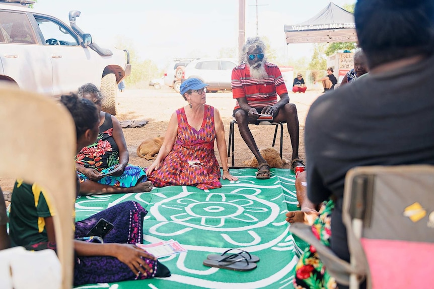 Lynne Walker sits on the ground on a rug and talks to locals in Mulka with four-wheel-drives in the background.