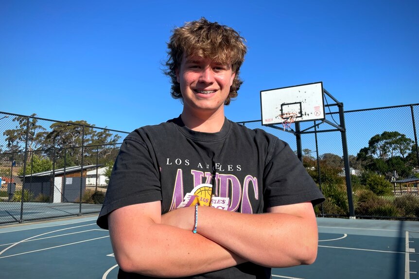 A young man with a blonde mullet in a navy coloured tshirt, arms crossed, standing on a basketball court smiling