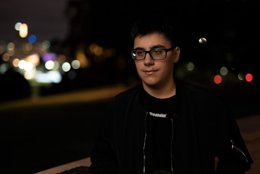 A teenage boy pictured at night, with blurred city lights behind him. He's looking off camera