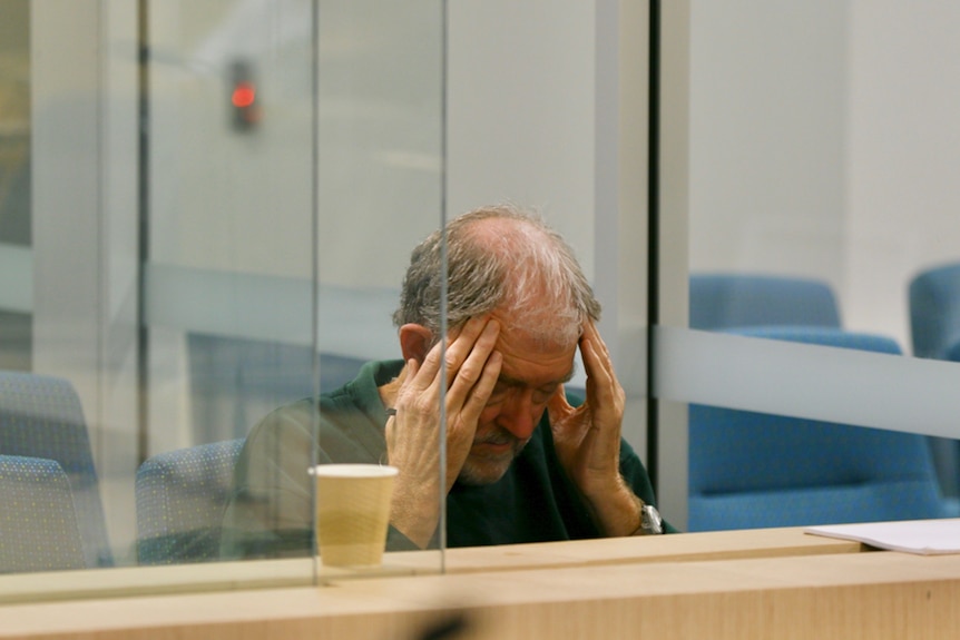 An older man with a bald head sits in a courtroom with his face in his hands.