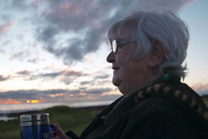 A woman holding a thermos mug watches the sunrise.