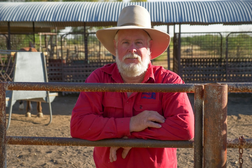 Man in red shirt and a hat leans on a fence in front of farm sheds.
