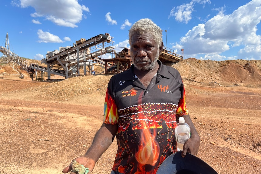 Indigenous man stands in front of old mine site with infrastructure left over