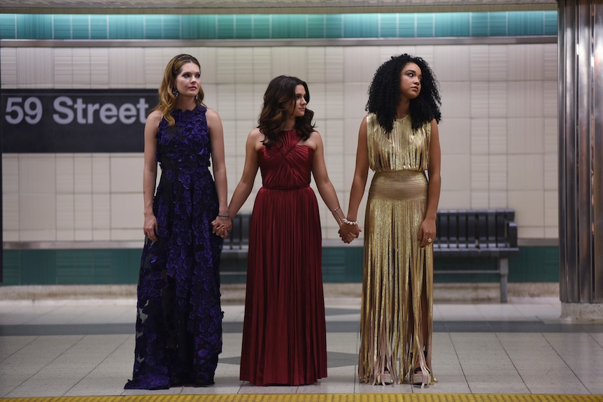 Three women in their 20s - two white and one Black - stand on a New York subway platform, holding hands and wearing fancy dress