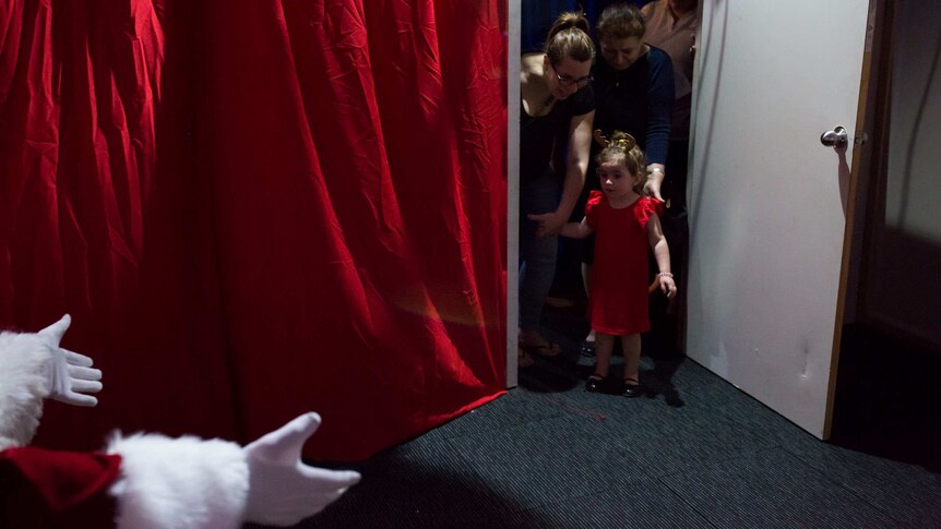 A little girl in a red dress stands timidly at the door as Santa's white-gloved hands reach for her.