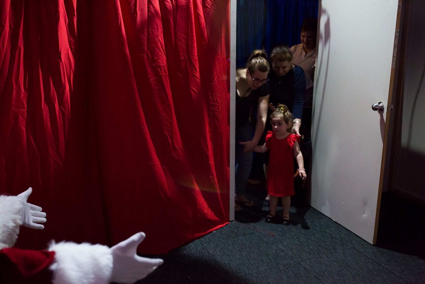 A little girl in a red dress stands timidly at the door as Santa's white-gloved hands reach for her.