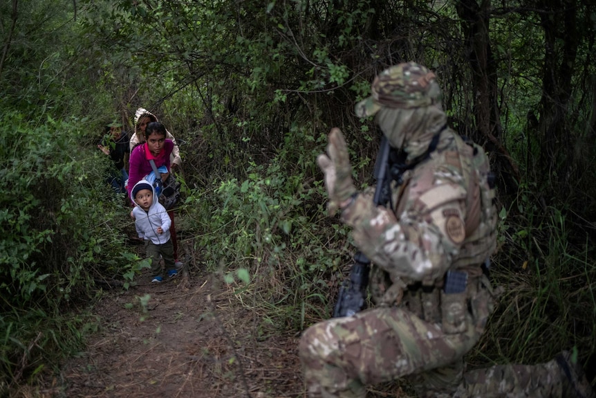 A man in heavy camouflage intercepts a family with young children in the woods