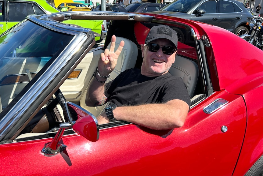 A smiling man flashes the peace sign as he sits in a vintage sports car.