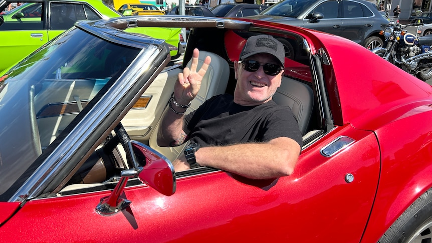 A smiling man flashes the peace sign as he sits in a vintage sports car.