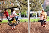 Children laugh and swing in a playground as a woman pushes one child on a swing.