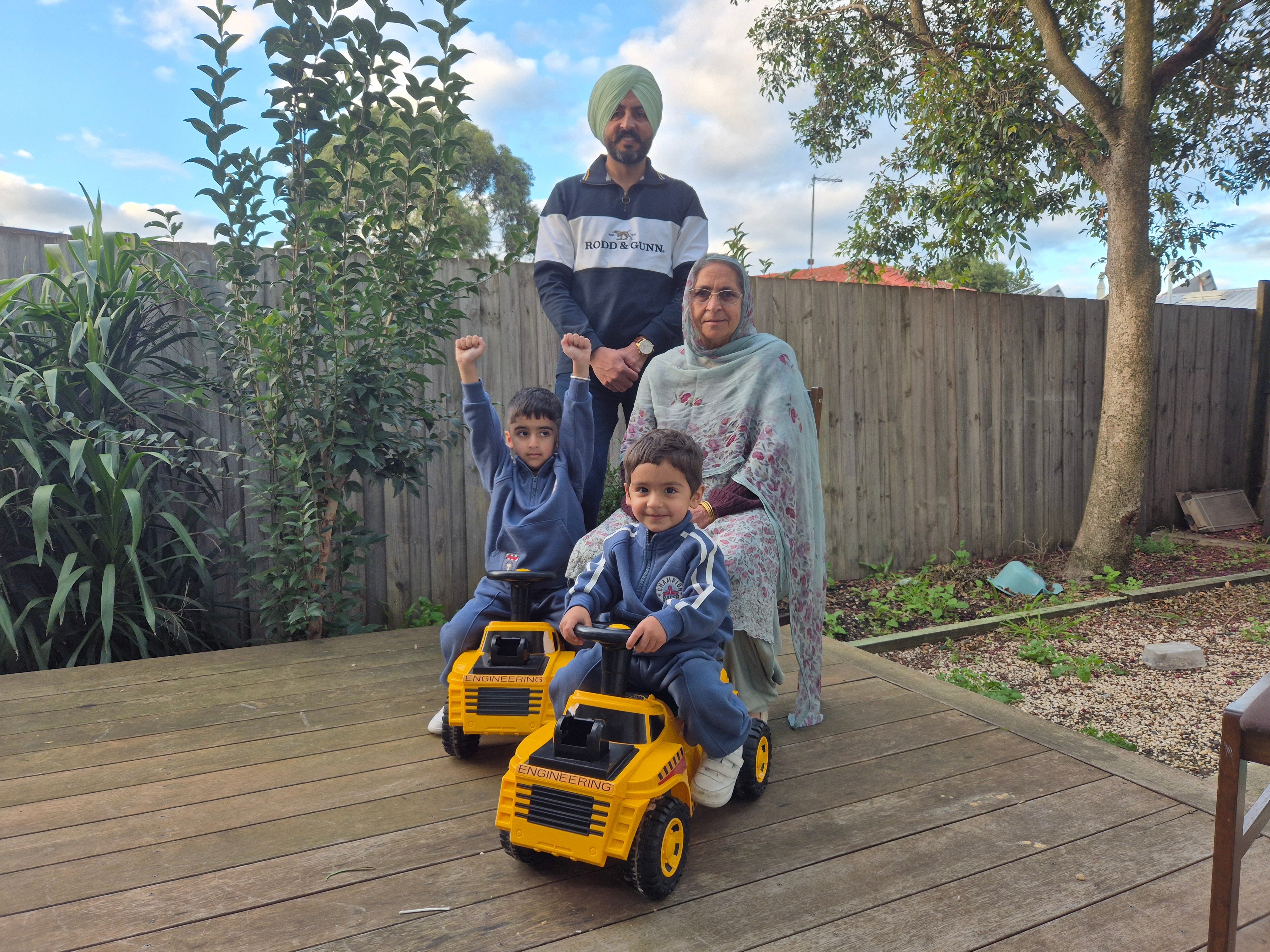 Satnam, Sikh man, wears  turban, smiles, stands behind mother, head covered with stole, two children on toy scooter, smile.
