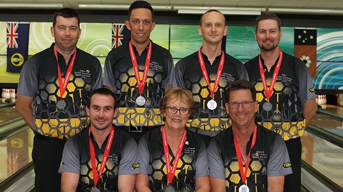 A bowling team stand in a bowling alley with medals around their necks.