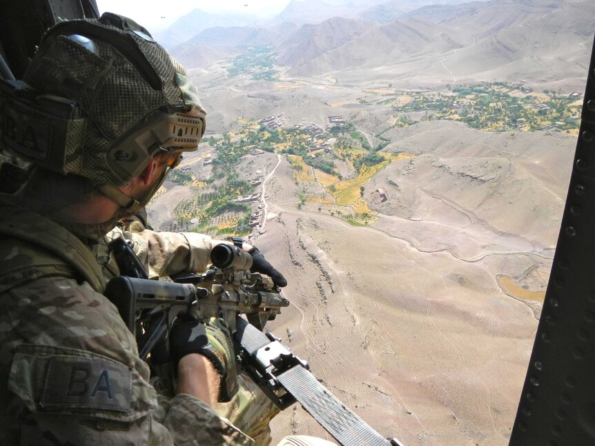 A soldier wearing a camouflage while holding a gun, looking out the side of a helicopter onto a brown mountainous area