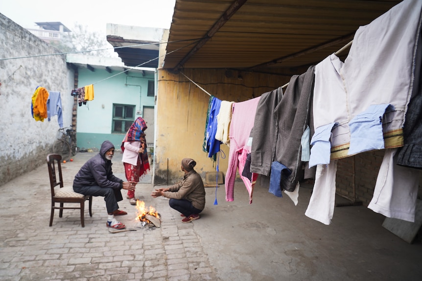 Three people sit outside their home next to a clothes line filled with clothes.