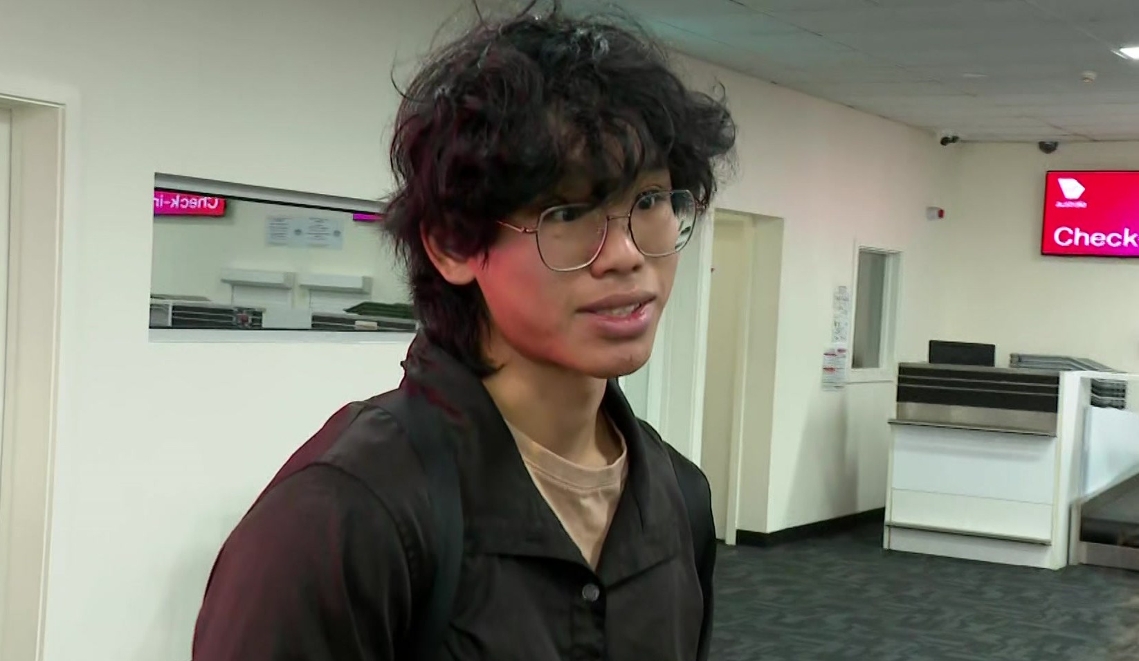A man with long wavy dark hair and glasses in a dark jacket and light brown shirt stares off camera.