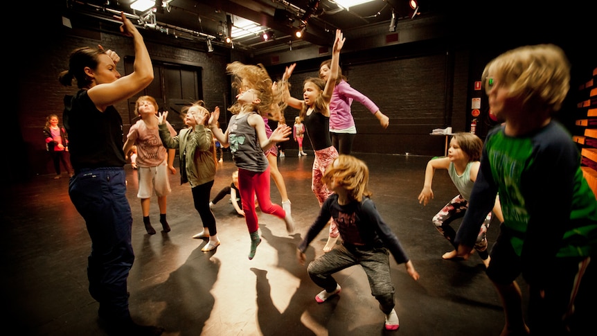 Group of children jumping and dancing in a black-painted studio, with an adult standing at front of them demonstrating.