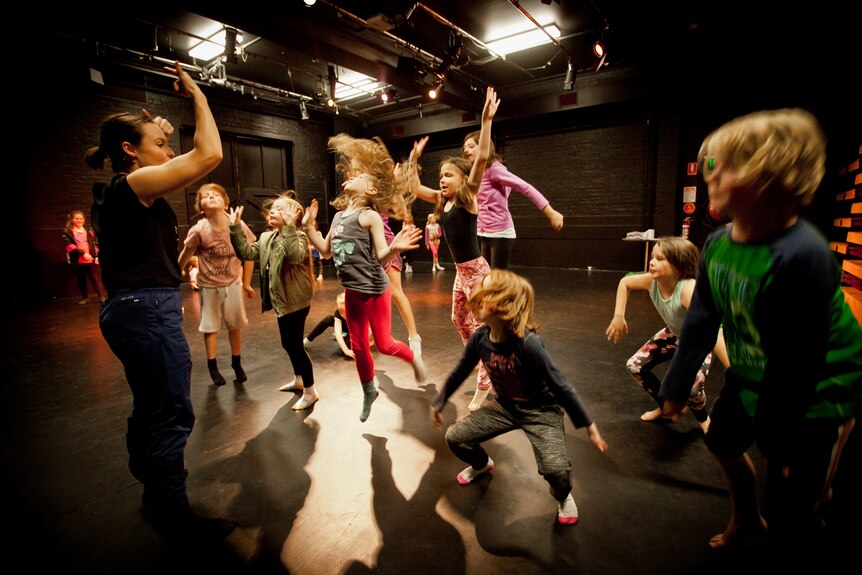 Group of children jumping and dancing in a black-painted studio, with an adult standing at front of them demonstrating.