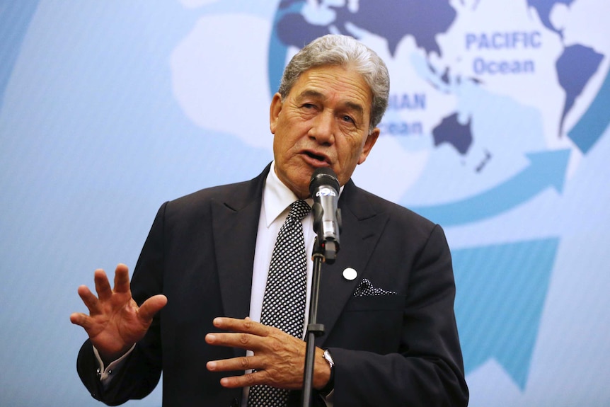 Wearing a black suit and tie, NZ Foreign Minister Winston Peters makes gestures with his hands as he speaks at a microphone.