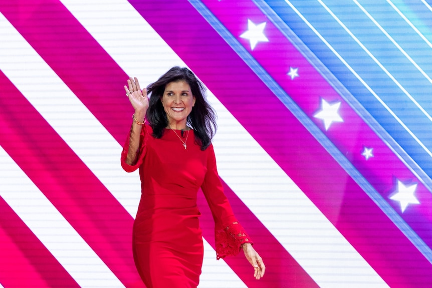 A woman waves as she walks in front of a billboard marked with stars and stripes