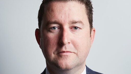 A head and shoulders photo of a man with short brown hair, wearing a blue suit jacket and tie.