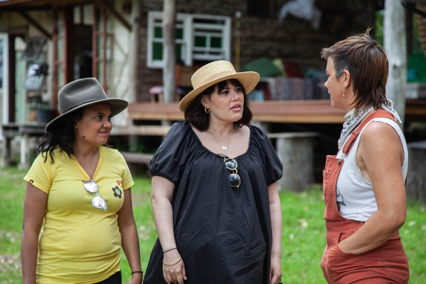 Miranda Tapsell and Nakkiah Lui in hats standing outside at farm, looking at Nina Pedersen who has overalls on, hands in pockets