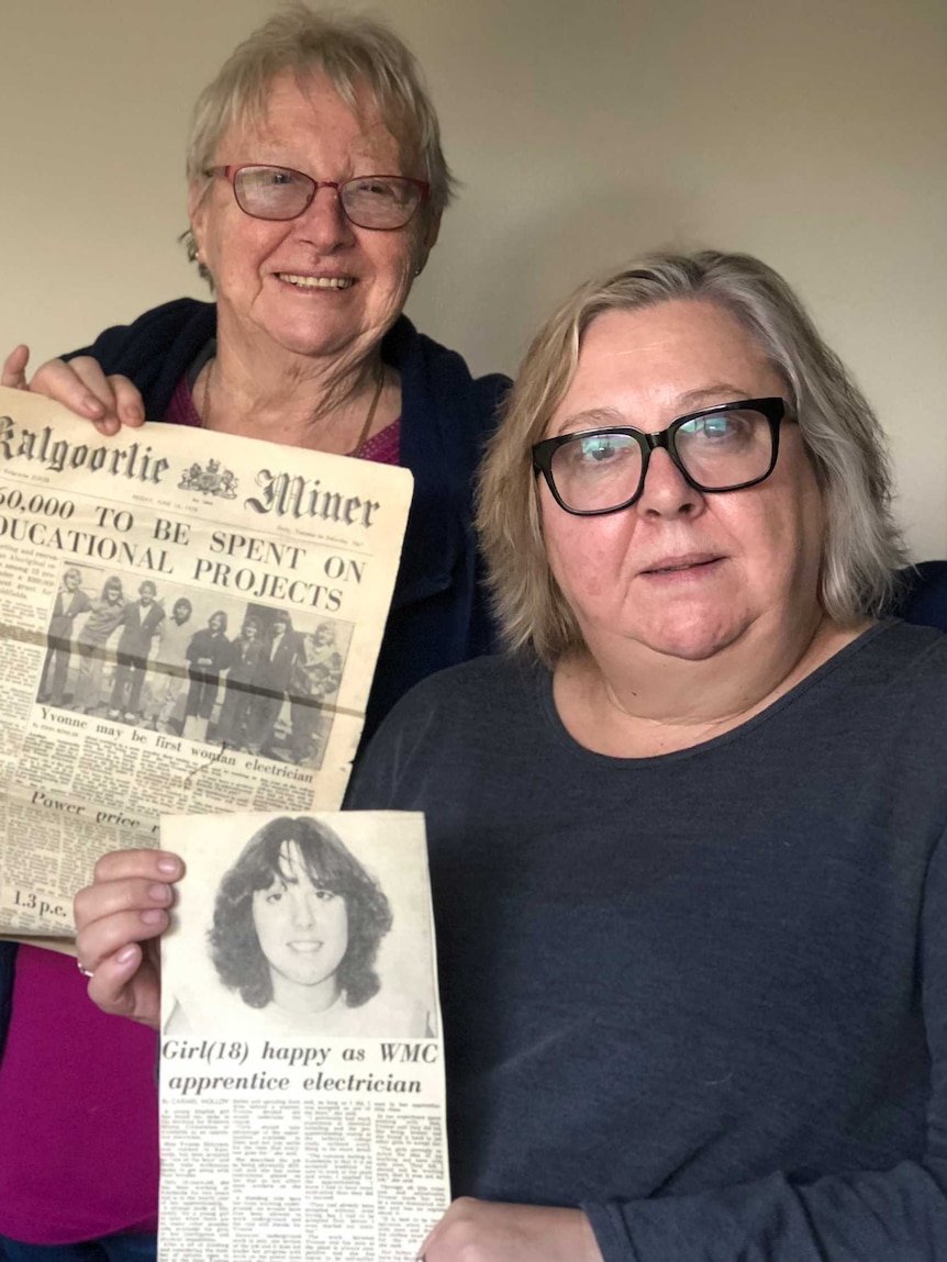 Two women in front of a blank wall holding up newspaper clippings